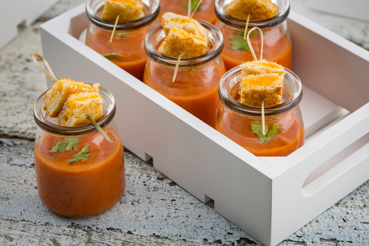 Small glass jars of tomato soup are topped with mini grilled cheese sandwiches, each held together by a bamboo pick with a looped end pick from TableCraft Products. Garnished with fresh herbs, they’re served on a white tray atop a rustic wooden surface.