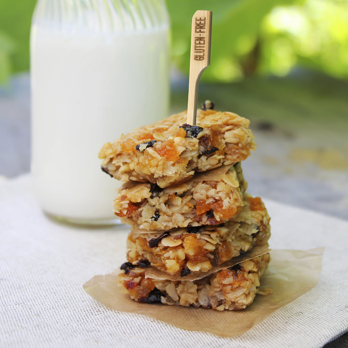 A stack of four oat and dried fruit bars sits on parchment paper, with a bamboo pick labeled “GLUTEN FREE” in the top bar. A glass bottle of milk is blurred in the background, and everything rests on a light cloth with green outdoor foliage behind.