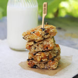 A stack of four oat and dried fruit bars sits on parchment paper, with a bamboo pick labeled “GLUTEN FREE” in the top bar. A glass bottle of milk is blurred in the background, and everything rests on a light cloth with green outdoor foliage behind.