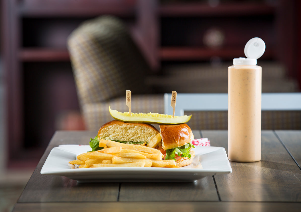 A sandwich with lettuce and a pickle slice on top is served on a white plate with French fries. Two bamboo sandwich picks hold the halves together. A squeeze bottle of orange sauce sits beside the plate on a wooden table, with a blurred background visible.