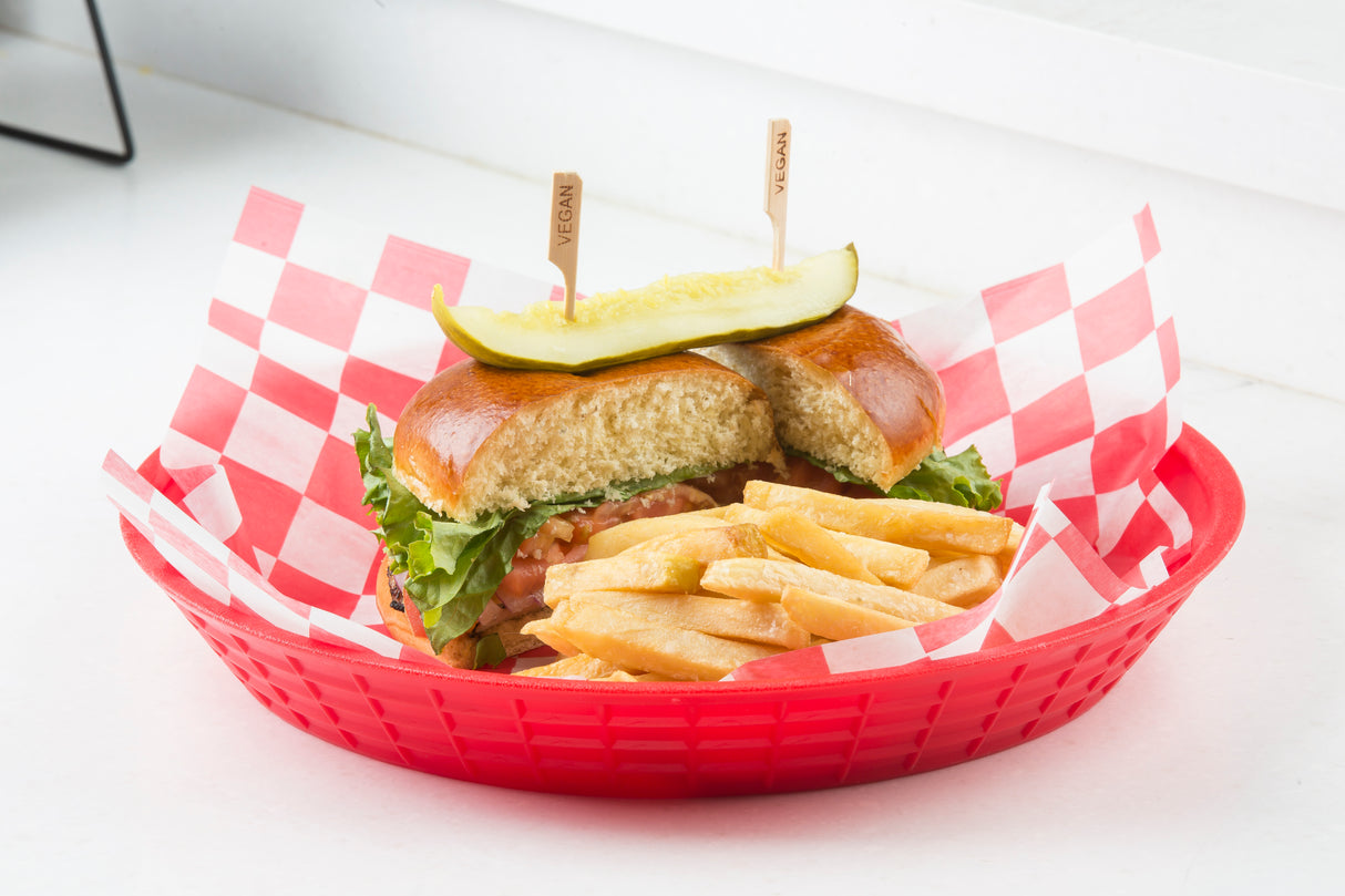 A red basket lined with red and white checkered paper holds a vegan sandwich topped with lettuce and a Vegan Pick, plus golden French fries. The sandwich is partially cut, revealing tomato and lettuce inside a soft bun.