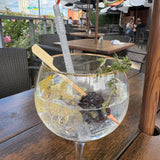 A large cocktail glass with ice, a dark fruit, herbs, a spiral straw, and a bamboo paddle pick sits on an outdoor table at a patio restaurant. Empty chairs and tables, pink flowers, and people dining are visible in the background.