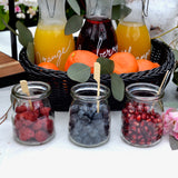 Three glass jars filled with raspberries, blueberries, and pomegranate seeds are in front of a black basket holding oranges and juice pitchers. TableCraft Products like a bamboo paddle pick add flair to this vibrant scene decorated with green leaves and a pink flower.