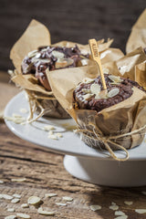 Three chocolate muffins sit on a white cake stand, wrapped in brown parchment and tied with twine. Topped with sliced almonds, one features a bamboo Allergy Pick from TableCraft Products. Almond slices are scattered on the rustic wooden surface.
