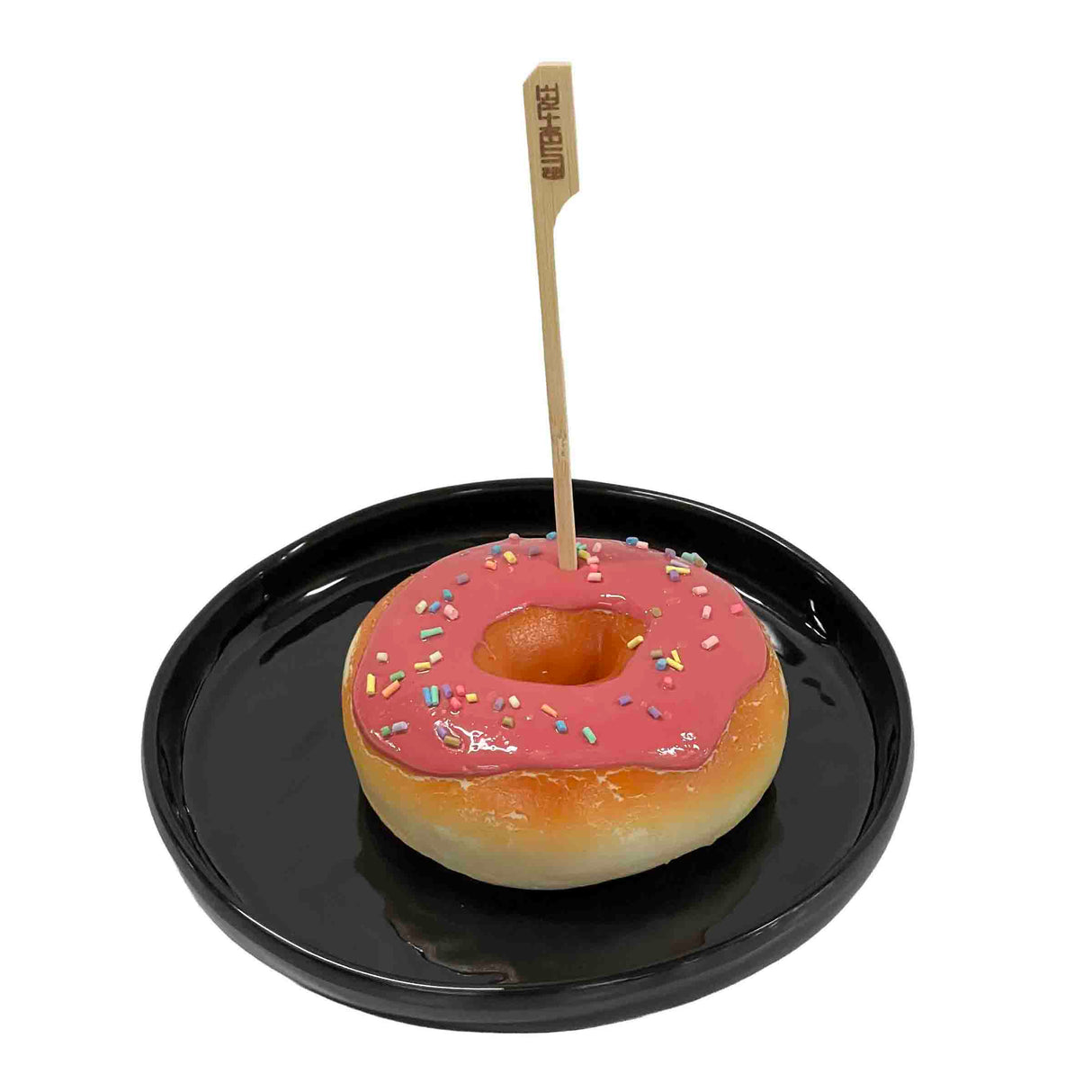 A round donut with pink icing and colorful sprinkles sits on a shiny black plate. A TableCraft Products Gluten-Free Pick, made of bamboo, is inserted vertically into the glossy donut’s center, clearly labeling it as gluten-free.