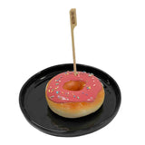 A round donut with pink icing and colorful sprinkles sits on a shiny black plate. A TableCraft Products Gluten-Free Pick, made of bamboo, is inserted vertically into the glossy donut’s center, clearly labeling it as gluten-free.