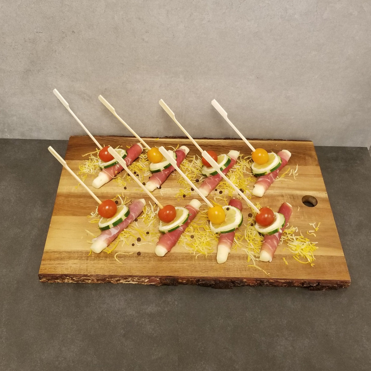 A wooden board displays eight toothpick appetizers made with white asparagus wrapped in prosciutto, topped with cucumber slices and cherry tomatoes, each secured with a 7 inch TableCraft bamboo paddle pick and placed on shredded cheese against a gray background.