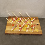 A wooden board displays eight toothpick appetizers made with white asparagus wrapped in prosciutto, topped with cucumber slices and cherry tomatoes, each secured with a 7 inch TableCraft bamboo paddle pick and placed on shredded cheese against a gray background.