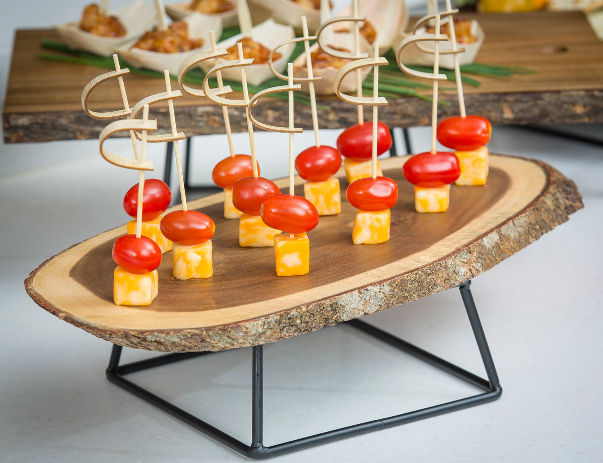 A rustic wooden serving tray displays skewers with marbled cheese and grape tomato, each held by a bamboo sword pick from TableCraft. The tray rests on a black metal stand, while other appetizers are slightly out of focus in the background.