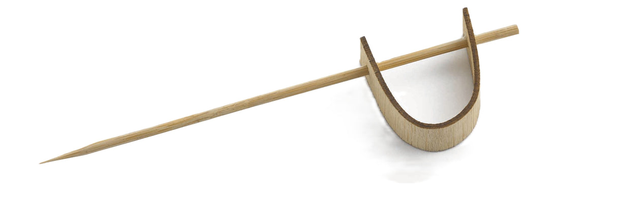 A TableCraft bamboo sword pick rests on a curved bamboo holder shaped like an open U. The pick is pointed at one end and lays diagonally across the holder, which sits on a white background. Perfect for cash and carry displays.