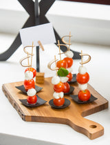 A wooden serving board holds five mini skewers with cherry tomatoes, mozzarella balls, and basil leaves on small black dishes. Each skewer features a bamboo sword pick. In the bright, minimalistic background is a white card clipped to a stand.