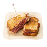 A Reuben sandwich cut in half sits on a wooden plate. Layers of corned beef, sauerkraut, and melted cheese fill toasted rye bread. One half is secured with a Sport Pick topped with a small baseball for a playful finish.