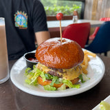 A cheeseburger with lettuce on a shiny bun is served on a white plate with fries and ketchup. The burger is topped with a sport pick featuring a small basketball. A milkshake and a person in a black shirt are in the background.