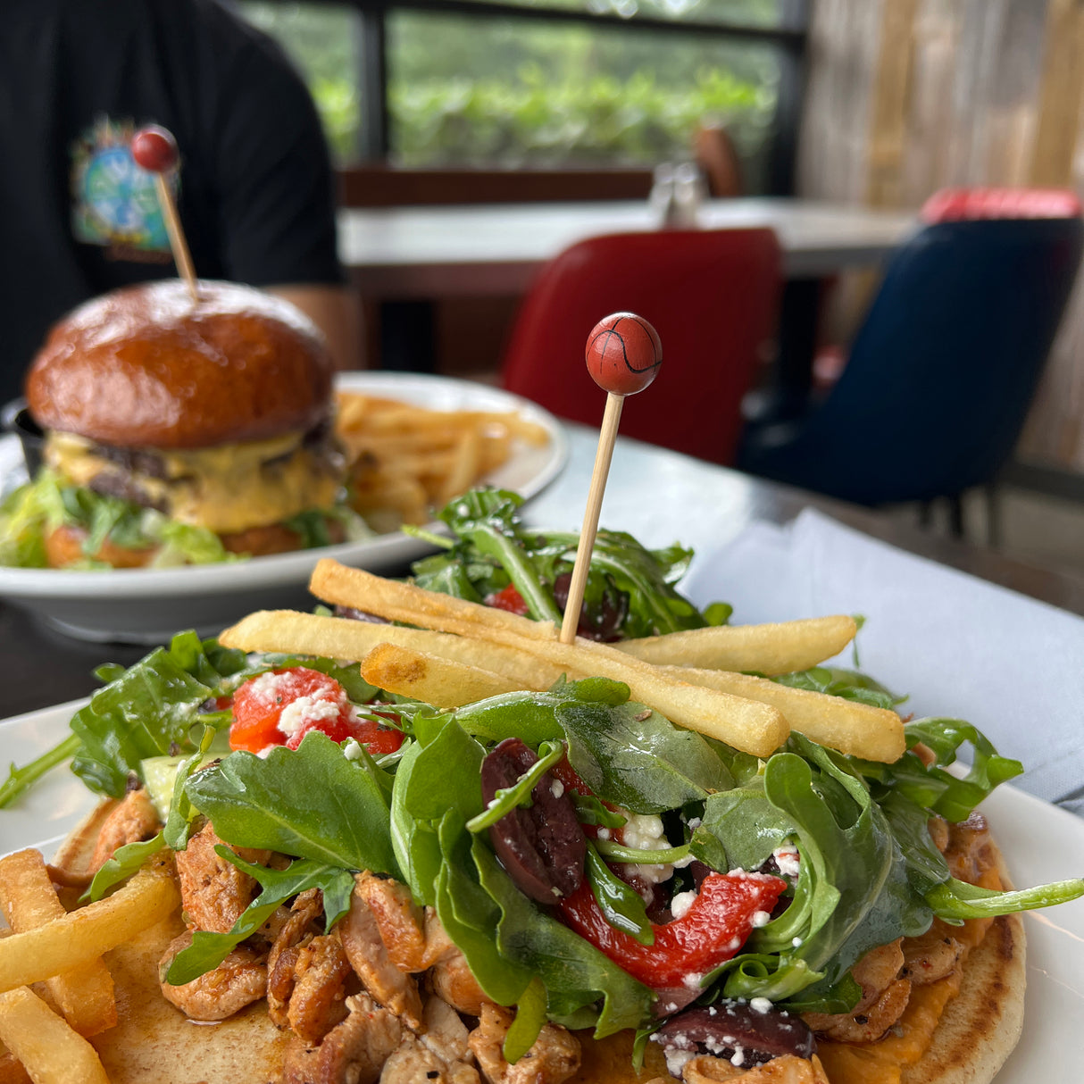 A close-up of a gyro topped with arugula, olives, red pepper, feta, and fries is served with more fries on the side and secured with a sport pick. In the background, a cheeseburger and fries sit near bamboo-accented indoor seating and colorful chairs.