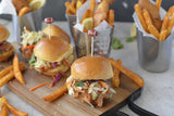 Two sliders with shredded coleslaw and sauce on brioche buns sit on a wooden board, each secured with a bamboo football pick. In the background, TableCraft Products metal cups hold fried fish and fries, garnished with lemon wedges for a fresh touch.