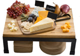 A collapsible wood butcher block cheese board on black legs holds grapes, figs, pickles, crackers, pears, and assorted cheeses—cheddar, blue, cubed, and wedge-shaped—some labeled with signs. Stacks of wooden plates and napkins sit underneath.