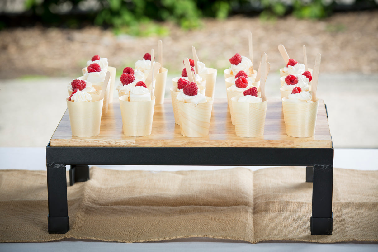 A collapsible wood butcher block riser on black metal legs displays twelve dessert cups with whipped cream and raspberries, each with a wooden spoon, arranged neatly on a table with a burlap runner. Blurred greenery decorates the background.
