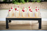 A collapsible wood butcher block riser on black metal legs displays twelve dessert cups with whipped cream and raspberries, each with a wooden spoon, arranged neatly on a table with a burlap runner. Blurred greenery decorates the background.