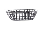 A black powder coated metal Serving Basket, oval-shaped with an open, grid-like pattern. Part of the Complexity Collection, the intersecting wires create a modern geometric design. The empty basket is set against a plain white background.
