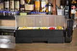 The TableCraft garnish organizer features a black bar fruit garnish tray with a clear lid holding lemons, limes, oranges, cherries, and olives. Cocktail straws sit to the right, with bottles and barware blurred in the background.