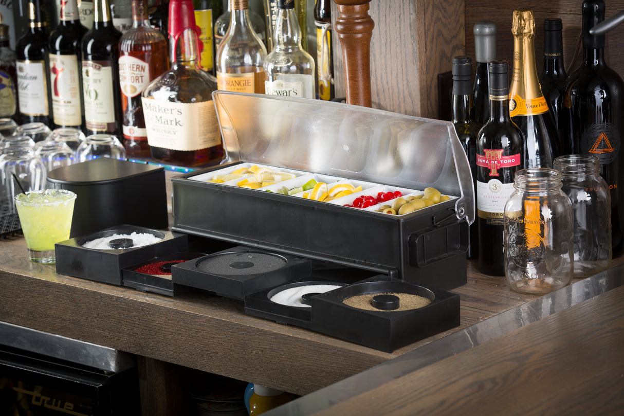 A bar counter with bottles of liquor and wine in the background. On the counter, a TableCraft garnish organizer holds sliced lemons, limes, cherries, and olives. Several open condiment containers are in front, alongside mason jars and a yellow cocktail.