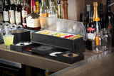 A bar counter with bottles of liquor and wine in the background. On the counter, a TableCraft garnish organizer holds sliced lemons, limes, cherries, and olives. Several open condiment containers are in front, alongside mason jars and a yellow cocktail.