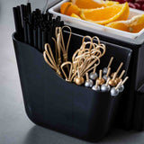 A TableCraft black organizer holds black plastic straws and decorative drink picks with metallic silver and gold beads. Behind the dishwasher safe organizer is a white tray filled with orange slices, sitting on a gray surface.