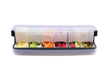 A transparent plastic garnish station with a lid features six compartments filled with lemon wedges, lime wedges, orange peel, cherries, strawberries, and green olives—all organized in a neat row for easy access.