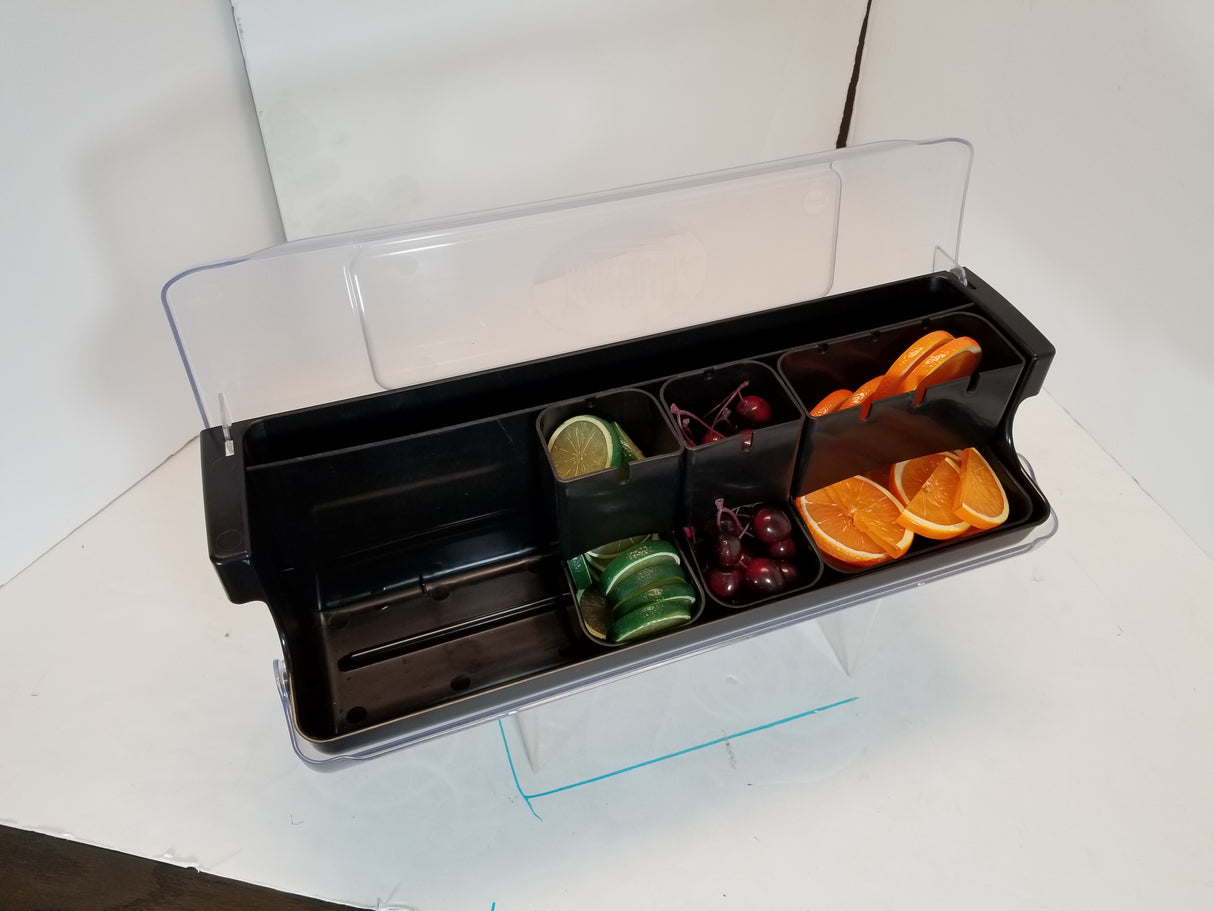 A black polypropylene bar garnish tray, model BCD6000, with a clear lid holds sliced limes, green olives, maraschino cherries, and orange slices in separate compartments on a white surface with a blue line beneath the tray.