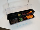 A black polypropylene bar garnish tray, model BCD6000, with a clear lid holds sliced limes, green olives, maraschino cherries, and orange slices in separate compartments on a white surface with a blue line beneath the tray.