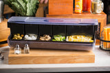 A plastic garnish station with five compartments holds green olives, pearl onions, mushrooms, lime wedges, and lemon wedges. Placed on a wooden board with salt and pepper shakers in front, it’s perfect for keeping bar supplies organized.