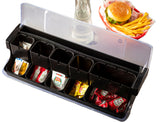 A condiment organizer with six compartments acts as a compact garnish station, holding single-serve ketchup, mustard, and relish packets. The clear-lidded NSF container sits beside a burger and French fries in a basket on a white background.
