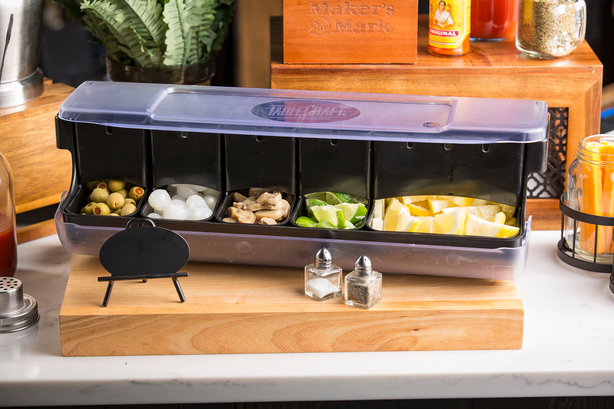 A bar condiment organizer made of dishwasher safe polypropylene on a wooden board holds green olives, cocktail onions, ice, mushrooms, lime and lemon wedges. Small salt and pepper shakers and a blank chalkboard sign sit in front; various bar items appear in the background.