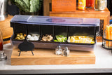 A bar condiment organizer made of dishwasher safe polypropylene on a wooden board holds green olives, cocktail onions, ice, mushrooms, lime and lemon wedges. Small salt and pepper shakers and a blank chalkboard sign sit in front; various bar items appear in the background.