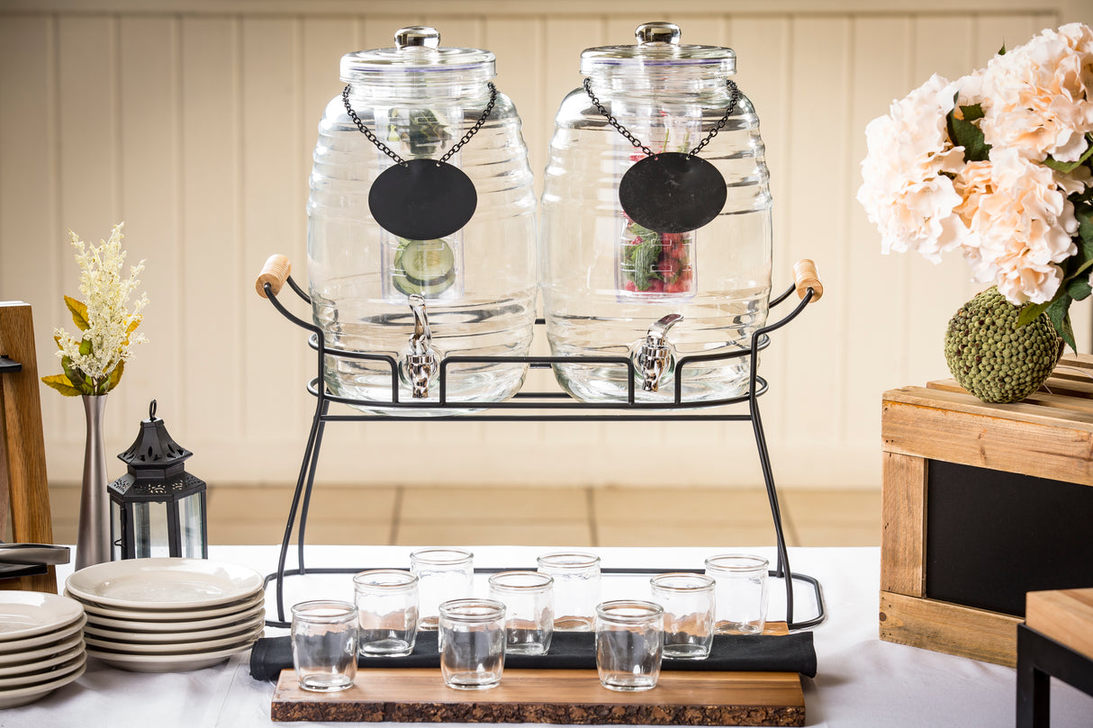 Two large TableCraft Products glass beverage dispensers with metal spigots sit on a black stand, each filled with water, lime, and berries. Eight empty glasses are arranged in front, with flowers, a lantern, and stacked plates decorating the table.