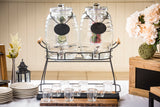 Two large TableCraft Products glass beverage dispensers with metal spigots sit on a black stand, each filled with water, lime, and berries. Eight empty glasses are arranged in front, with flowers, a lantern, and stacked plates decorating the table.