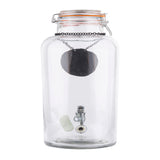 A clear glass beverage dispenser with a hinged lid contains a black pendant necklace hanging from the top. Inside, at the bottom, are a small white cylindrical object, a metal clasp, and an artificial eyeball. The background is plain white.