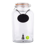 A rustic 2.5 gallon glass beverage dispenser with a spigot at the bottom center, an orange-sealed hinged lid, and a blank black chalkboard label on a chain. A small green sticker sits near the base on the front.