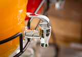 A close-up of a shiny metal replacement faucet attached to a bright orange glass beverage dispenser, with a red dispenser blurred in the background. The spigot is clean and reflective, and the TableCraft dispenser rests on a black metal wire stand.