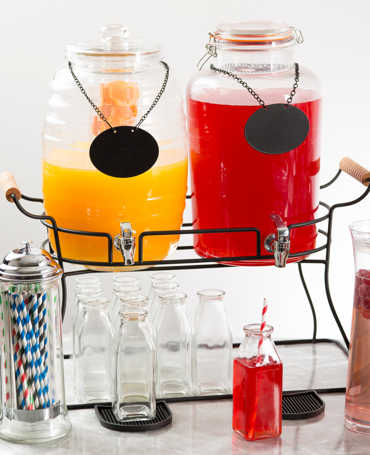 Two large double beverage dispensers, one with orange juice and one with red punch, sit on a black powder coated metal glass beverage dispenser base. Empty glass bottles, striped straws, and a tall fruit-garnished glass are in front. Both have blank black labels hanging from chains.