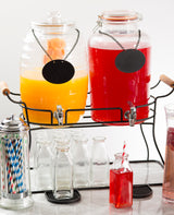 Two large double beverage dispensers, one with orange juice and one with red punch, sit on a black powder coated metal glass beverage dispenser base. Empty glass bottles, striped straws, and a tall fruit-garnished glass are in front. Both have blank black labels hanging from chains.