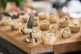 Several small pinwheel sandwiches with visible fillings of spinach and cheese are arranged on a wooden serving board. A stainless steel fork clip is stuck upright in one sandwich, resembling a number card holder in this casual dining scene.