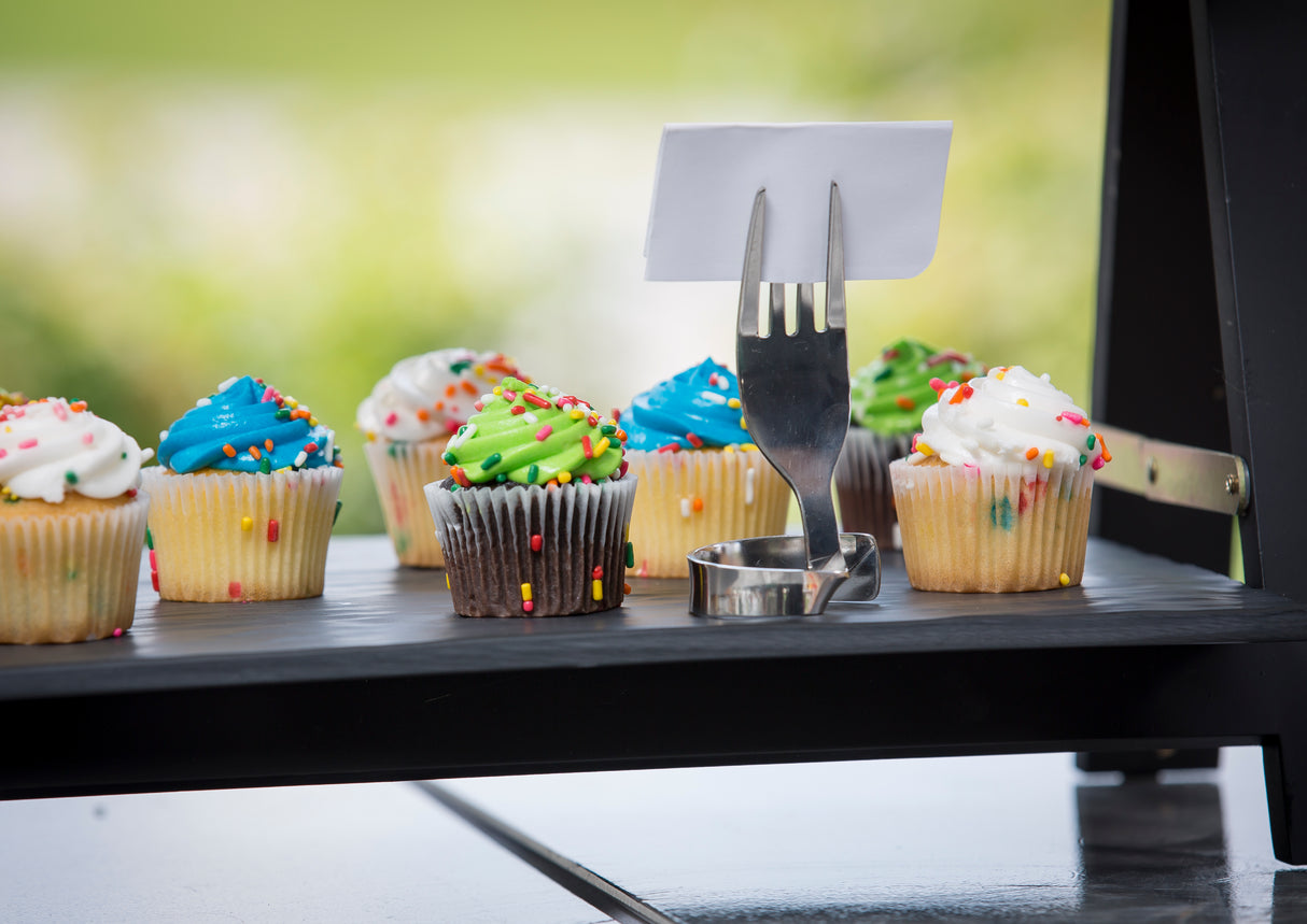 Colorful cupcakes with sprinkles are arranged on a black tray. A stainless steel fork clip serves as a number card holder, displaying a blank white card upright in the center. The blurred background of greenery hints at an outdoor setting.