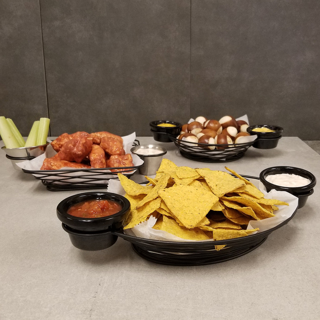A table with trays of snacks: tortilla chips with salsa and sour cream, saucy chicken wings with celery and ranch dip, and soft pretzel bites with mustard. The black powder coated metal trays feature integrated ramekin holders for easy dipping.