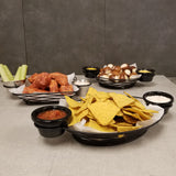 A table with trays of snacks: tortilla chips with salsa and sour cream, saucy chicken wings with celery and ranch dip, and soft pretzel bites with mustard. The black powder coated metal trays feature integrated ramekin holders for easy dipping.