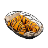 A black powder coated metal Artisan Collection Basket, oval in shape and lined with beige cloth, holds three golden-brown croissants drizzled with dark chocolate on a white background.