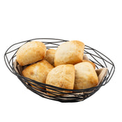 A black powder coated metal basket from the Artisan Collection Basket, lined with beige cloth, holds several golden-brown, freshly baked bread rolls with rustic exteriors, arranged close together on a white background.