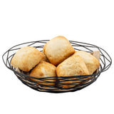 A black powder coated metal basket from the Artisan Collection Basket line, model TableCraft BK17412, holds six golden-brown, rustic dinner rolls piled atop a beige cloth, all isolated on a white background.