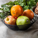 A black powder coated metal fruit bowl from the TableCraft Artisan Collection Basket holds two oranges, a green pear, and a red apple on a gray surface. Green leafy plants and part of a seeded bread roll blur in the background. Natural light highlights the fresh fruit.