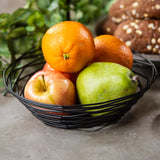 A black powder coated metal Artisan Collection Basket by TableCraft holds a green pear, a red apple, and two oranges. In the blurred background, leafy greens and oat-topped bread rolls sit on a light gray surface.